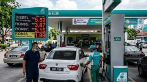 A realistic photograph capturing a fuel attendant in a Petronas uniform refueling a white Proton Saga car (plate BND 2026) at a busy Malaysian petrol station. A prominent digital price board displays fuel prices including Diesel at RM 5.52 and subsidized RON95 at RM 1.99, alongside a 'BUDI MADANI' sign.