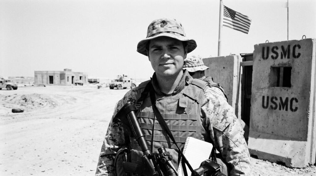A 16:9 black and white photo of young JD Vance in U.S. Marine Corps combat gear and military hat during his deployment to Iraq.