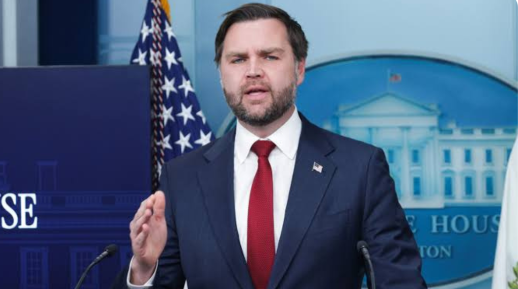 A full view of JD Vance as the Vice President of the United States, in a blue suit with a red tie, speaking confidently at a press conference podium in the White House briefing room, flanked by US flags.