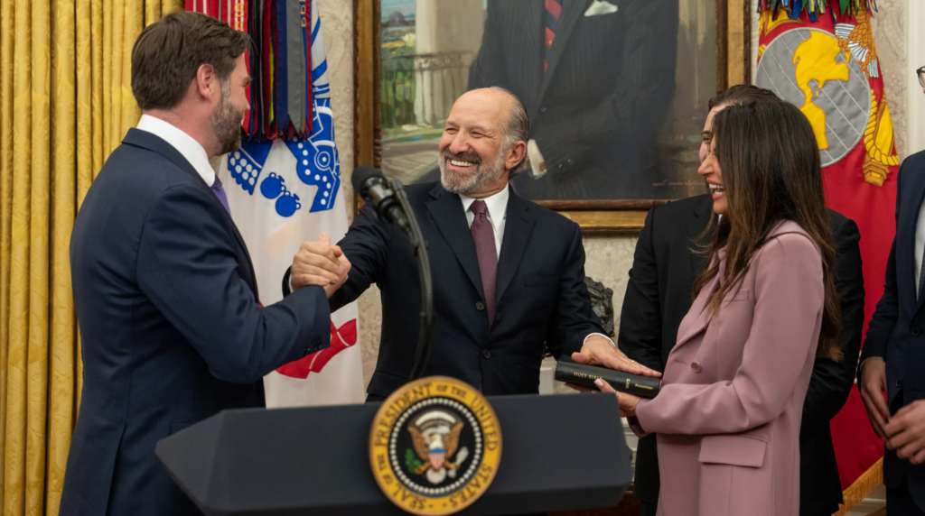 Howard Lutnick, smiling, shakes hands with Vice President JD Vance while Allison Lambert holds a Bible for his swearing-in as Secretary of Commerce in the Oval Office.