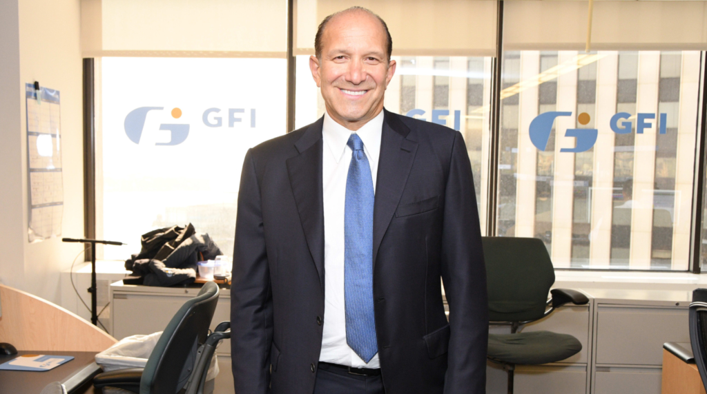 Howard Lutnick, long-time CEO of Cantor Fitzgerald, standing in a corporate office wearing a dark suit and blue tie.